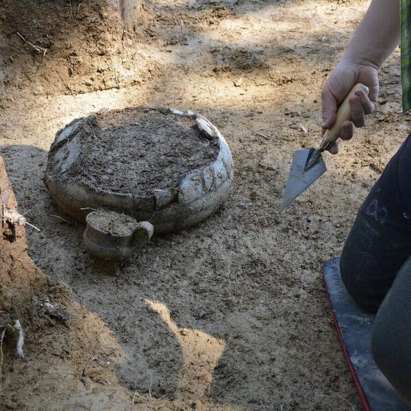 Eine Person verwendet eine kleine Kelle, um an einer archäologischen Ausgrabungsstätte teilweise im Boden vergrabene Keramikstücke auszugraben.