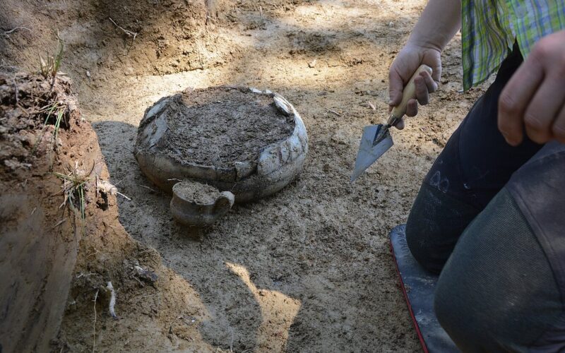 Eine Person verwendet eine kleine Kelle, um an einer archäologischen Ausgrabungsstätte teilweise im Boden vergrabene Keramikstücke auszugraben.
