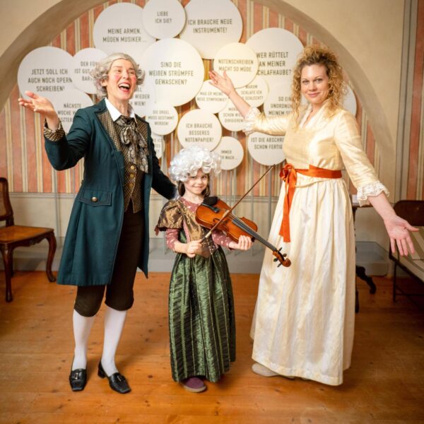Drei Personen in historischen Kostümen, ein Kind mit einer Geige, posieren und lächeln drinnen vor einer Wand mit deutschen Sprechblasen.
