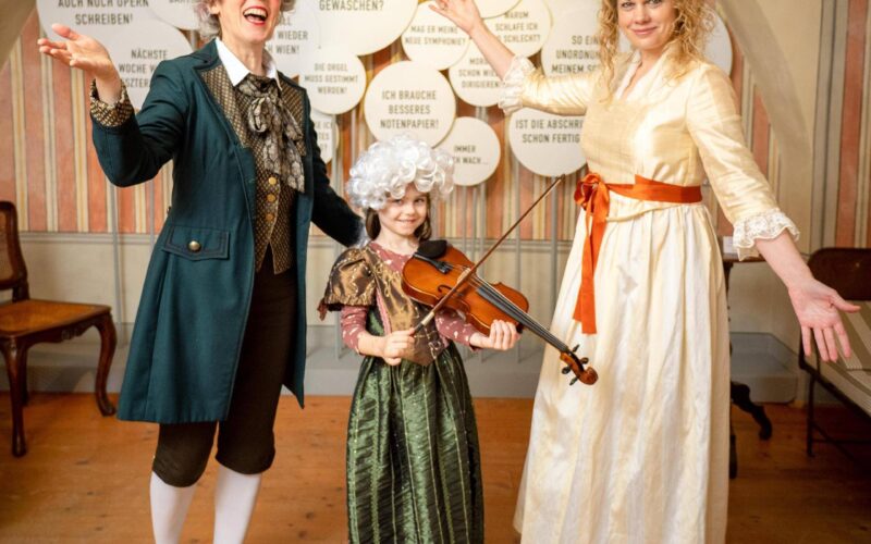 Drei Personen in historischen Kostümen, ein Kind mit einer Geige, posieren und lächeln drinnen vor einer Wand mit deutschen Sprechblasen.