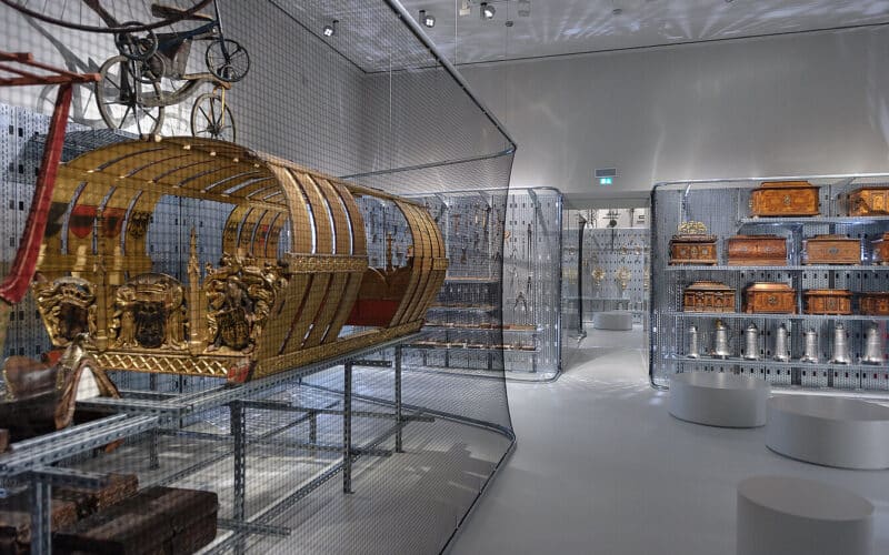 Ausstellungsraum des Museums mit kunstvollen Kutschen, Truhen und Dekorationsgegenständen in Drahtgittervitrinen unter heller Beleuchtung.