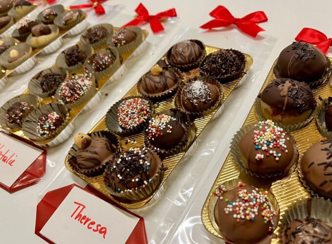 Tabletts mit verschiedenen Schokoladentrüffeln und verschiedenen Toppings, einzeln in goldenen Tabletts platziert und mit roten Namensschildern und Schleifen beschriftet.