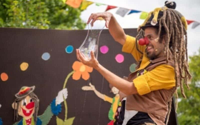 Ein als Clown verkleideter Darsteller mit roter Nase und Dreadlocks dehnt eine durchsichtige, elastische Substanz auf einer mit bunten Kunstwerken und Fahnen geschmückten Außenbühne.