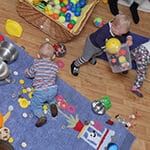 Drei Kleinkinder spielen auf einem Holzboden, auf dem verschiedene Spielzeuge, Bälle und Küchenutensilien verstreut sind; zwei befinden sich auf einer blauen Matte, eines in der Nähe eines Korbes mit Bällen.