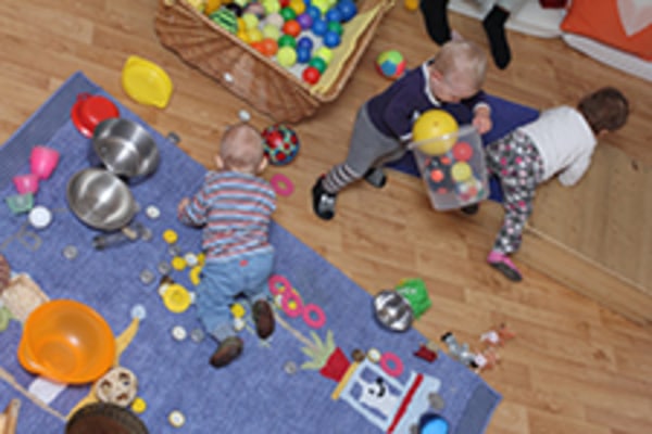 Drei Kleinkinder spielen drinnen auf einem Holzboden mit verschiedenen Spielsachen, Bällen und Küchenutensilien, die um eine Matte und einen Korb mit bunten Bällen herum verstreut sind.