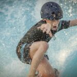 Ein Kind, das einen Helm und Badekleidung trägt, fährt auf einem Surfbrett auf blauem Wasser und hebt einen Arm, um das Gleichgewicht zu halten, während das Wasser herumspritzt.