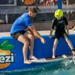 Ein Erwachsener hilft einem Kind in einem Helm mit einem Wakeboard auf einer künstlichen Welle, auf der ein "Spezi"-Logo an der Seite sichtbar ist.