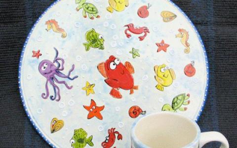 Ein Keramikteller und eine Tasse mit bunten Cartoon-Meerestieren, darunter Fische, Schildkröten, ein Oktopus und Seesterne, auf hellem Hintergrund.