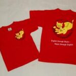 Zwei rote T-Shirts mit einer gelben Katzengrafik; ein Shirt hat die große Grafik und den Text "English through Music - Music through English", das andere hat ein kleines Brustlogo.