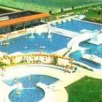 Luftaufnahme eines öffentlichen Freibadkomplexes mit mehreren Becken, einer Rutsche, einer Brücke, grünen Rasenflächen und einem nahe gelegenen Gebäude.