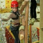 Eine Person, die einen roten Helm und einen Sicherheitsgurt trägt, überquert ein Hindernis aus Baumstämmen in einem Indoor-Hochseilgarten, während im Hintergrund die Kletterwände zu sehen sind.