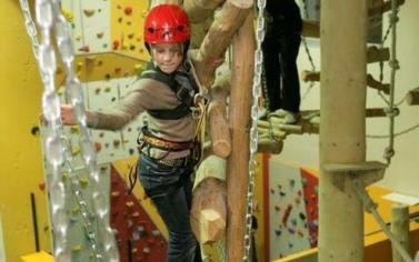Eine Person, die einen roten Helm und einen Sicherheitsgurt trägt, überquert ein Hindernis aus Baumstämmen in einem Indoor-Hochseilgarten, während im Hintergrund die Kletterwände zu sehen sind.