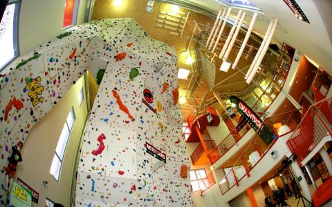 Indoor-Kletterhalle mit einer hohen, mehrfarbigen künstlichen Kletterwand, roten Geländern und einer Treppe in einem hell erleuchteten Raum.