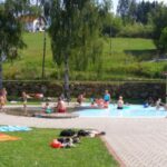 An einem sonnigen Tag entspannen sich die Menschen im Gras und schwimmen in einem kleinen Freibad, während die Kinder im und am Wasser spielen.