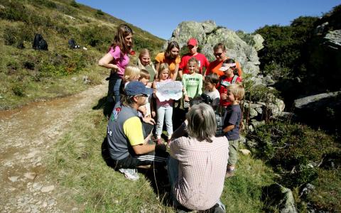 Eine Gruppe von Kindern und Erwachsenen versammelt sich im Freien auf einem grasbewachsenen Hügel und hört einem sitzenden Lehrer zu, der ein Whiteboard hält.