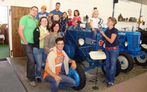 Eine Gruppe von Menschen, darunter Kinder und Erwachsene, posieren gemeinsam um einen blauen Oldtimer-Traktor in einem Innenraum. Einige Kinder sitzen auf dem Traktor, während andere in der Nähe stehen.