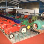 Eine Sammlung alter Traktoren und Maschinen, die drinnen auf einem roten Teppich ausgestellt sind, mit Industriebeleuchtung und Plastikstühlen in der Nähe.