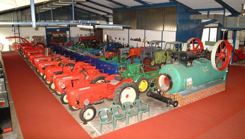Eine Sammlung alter Traktoren und Maschinen, die drinnen auf einem roten Teppich ausgestellt sind, mit Industriebeleuchtung und Plastikstühlen in der Nähe.