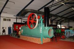 Eine große alte Dampfmaschine mit rotem Schwungrad steht in einem Innenraum auf einer gemauerten Plattform, umgeben von Industrieanlagen.