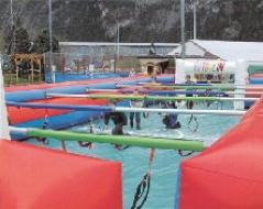 Auf einem aufblasbaren Spielfeld im Freien spielen Menschen Tischfußball, die jeweils an Schiebestangen befestigt sind, während im Hintergrund Berge und Gebäude zu sehen sind.