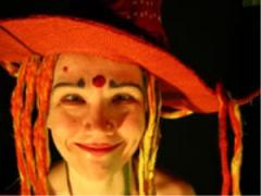 Lächelnde Person, die einen großen roten Hut mit Garn- oder Stofffäden und bunter Gesichtsbemalung trägt, darunter ein roter Punkt auf der Stirn und blaue Akzente über den Augen.