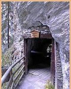 Hölzerner Eingang zu einem engen Tunnel, der in einen Felsen gebaut ist, mit einem Holzgeländer auf der linken Seite und einem kleinen Schild über dem Eingang.