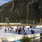 Menschen laufen Schlittschuh auf einer von einem Zaun umgebenen Eisbahn im Freien, im Hintergrund sind Berge und Bäume zu sehen.