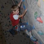 Ein Junge klettert mit einem Klettergurt an einer Indoor-Kletterwand und hält sich an bunten Griffen fest.