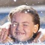 Drei Kinder mit nassen Haaren lächeln und halten sich gegenseitig in einem Schwimmbad, während das Wasser um sie herum spritzt.