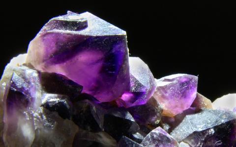 Nahaufnahme einer Gruppe violetter Amethystkristalle, die scharfe Kanten und durchscheinende Oberflächen vor einem dunklen Hintergrund zeigt.