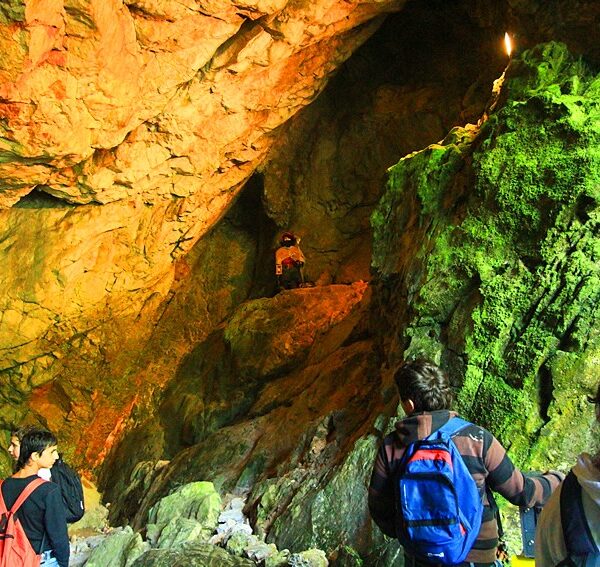 Eine Gruppe von Menschen mit Rucksäcken erkundet eine Höhle mit felsigen Wänden, orangefarbener Beleuchtung und grünen, moosbewachsenen Oberflächen.