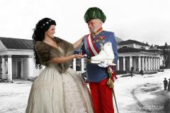 Eine Frau in einem historischen Kleid gestikuliert in Richtung eines Mannes in einem Kostüm mit Orden und Federhut, der einen modernen weißen Stiefel vor einem schwarz-weißen Hintergrund hält.