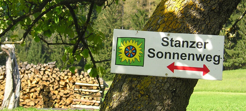 Ein Schild mit der Aufschrift "Stanzer Sonnenweg" mit einem Sonnensymbol und einem roten Pfeil ist an einem Baum auf einer Wiese in der Nähe von gestapeltem Brennholz angebracht.