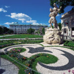 Ein formaler Garten mit dekorativ gemusterten Blumenbeeten, einer großen Steinstatue und einem historischen Gebäude im Hintergrund unter blauem Himmel.