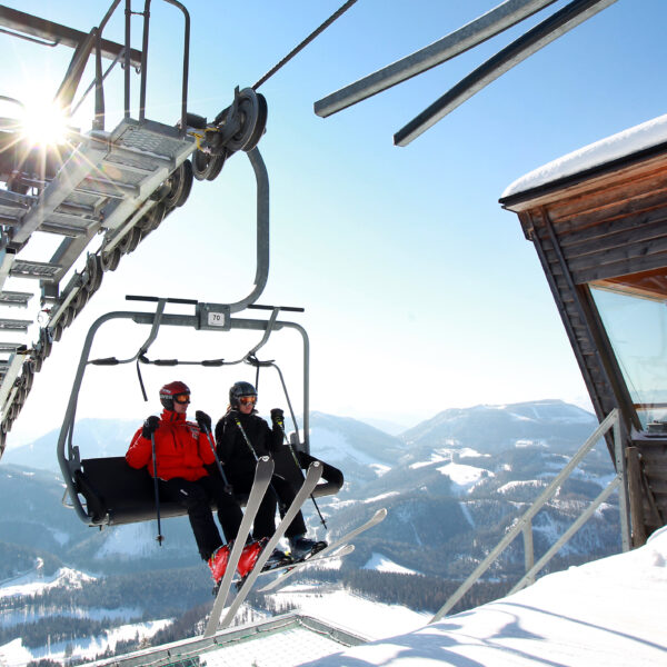 Zwei Skifahrer fahren mit einem Sessellift über verschneite Berge, vorbei an einer hölzernen Kabine des Liftbetreibers und unter strahlend blauem Himmel.