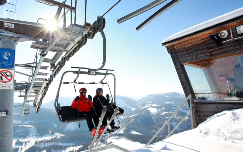 Zwei Skifahrer fahren mit einem Sessellift über verschneite Berge, vorbei an einer hölzernen Kabine des Liftbetreibers und unter strahlend blauem Himmel.