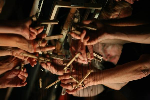 Mehrere Hände manipulieren an Schnüren befestigte Holzstäbe, was an die Steuerung von Marionetten erinnert. Die Szene ist schwach beleuchtet und konzentriert sich auf die koordinierte Bewegung.