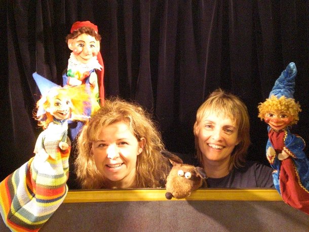 Zwei Frauen stehen hinter einem Puppentheatervorhang, jede hält bunte Handpuppen, vor einem schwarzen Stoffhintergrund.