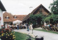 Vor einem rustikalen Gebäude mit Ziegeldach sind Menschen an Tischen im Freien versammelt, umgeben von Pflanzen und Blumen. Im Vordergrund ist ein Fahrrad geparkt.