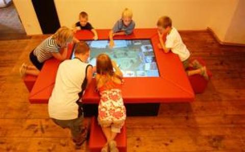 Sechs Kinder interagieren mit einem großen Touchscreen-Tisch in einem Raum mit Holzfußboden, wobei jedes von ihnen auf roten Sitzen um den Tisch herum sitzt oder sich anlehnt.