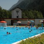 Freibad mit schwimmenden und spielenden Menschen; Berge und Bäume im Hintergrund unter einem klaren Himmel.