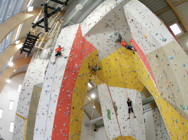 In einer geräumigen Kletterhalle klettern mehrere Personen an hohen Indoor-Felswänden mit verschiedenfarbigen Griffen.