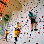 Vier Kinder in Klettergurten klettern an einer Indoor-Kletterwand in einer Turnhalle mit orangefarbenen Böden und Wänden.