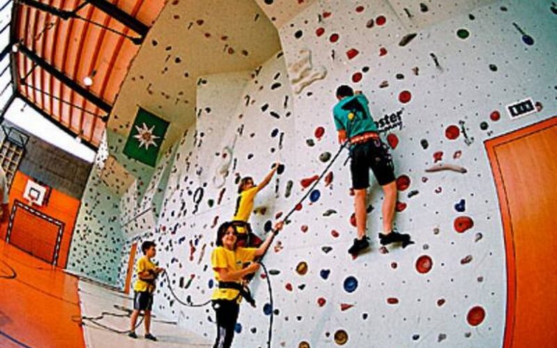Vier Kinder in Klettergurten klettern an einer Indoor-Kletterwand in einer Turnhalle mit orangefarbenen Böden und Wänden.