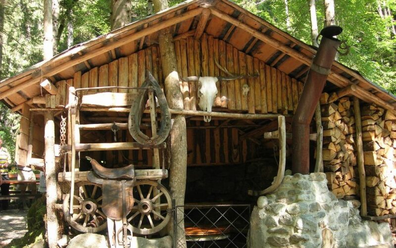 Eine rustikale Holzhütte mit gestapeltem Brennholz, einem montierten Rinderschädel, einem alten Wagenrad und einem Sattel an der Außenwand.