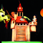 Eine Person bedient eine Drachenpuppe über einer Burgkulisse mit einer Puppenfigur auf einem Türmchen und einem orangefarbenen Kreis, der die Sonne im Hintergrund darstellt.