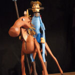 Eine hölzerne Puppenfigur eines Mannes mit Hut, der auf einem Pferd reitet und eine Lanze hält, vor einem dunklen Hintergrund.