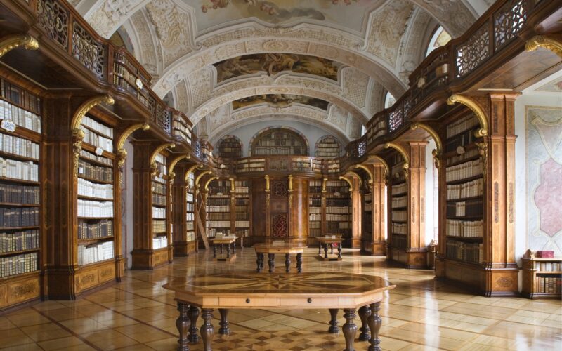 Eine große historische Bibliothek mit verzierten Gewölbedecken, hohen Bücherregalen und einem großen Holztisch in der Mitte.