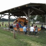 Die Menschen sitzen und stehen unter einem hölzernen Unterstand mit einer aufblasbaren Dinosaurierrutsche in einem grasbewachsenen Außenbereich.
