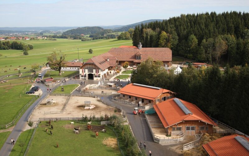 Luftaufnahme eines ländlichen Bauernhofs mit mehreren rotgedeckten Gebäuden, eingezäunten Tiergehegen, grünen Feldern und bewaldeten Flächen im Hintergrund.